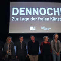Gruppenbild: Applaus für das Film-Team um die Regisseurin Janina Möbius vor einem Tableau mit dem Titel "DENNOCH! Zur Lage der freien Künste". Im Hintergrund ist Holger Bergmann mit einem Mikrofon in der Hand zu sehen.