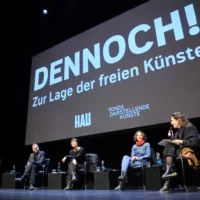 Panel discussion after the film screening with guests from politics and art. They sit in a row of chairs on the stage, above them is a projection with the title of the film.