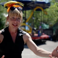 With open arms, an artist speaks expressively into the camera, a sun and an artificial bird in her hair. In the background of the picture a carousel is dimly visible.