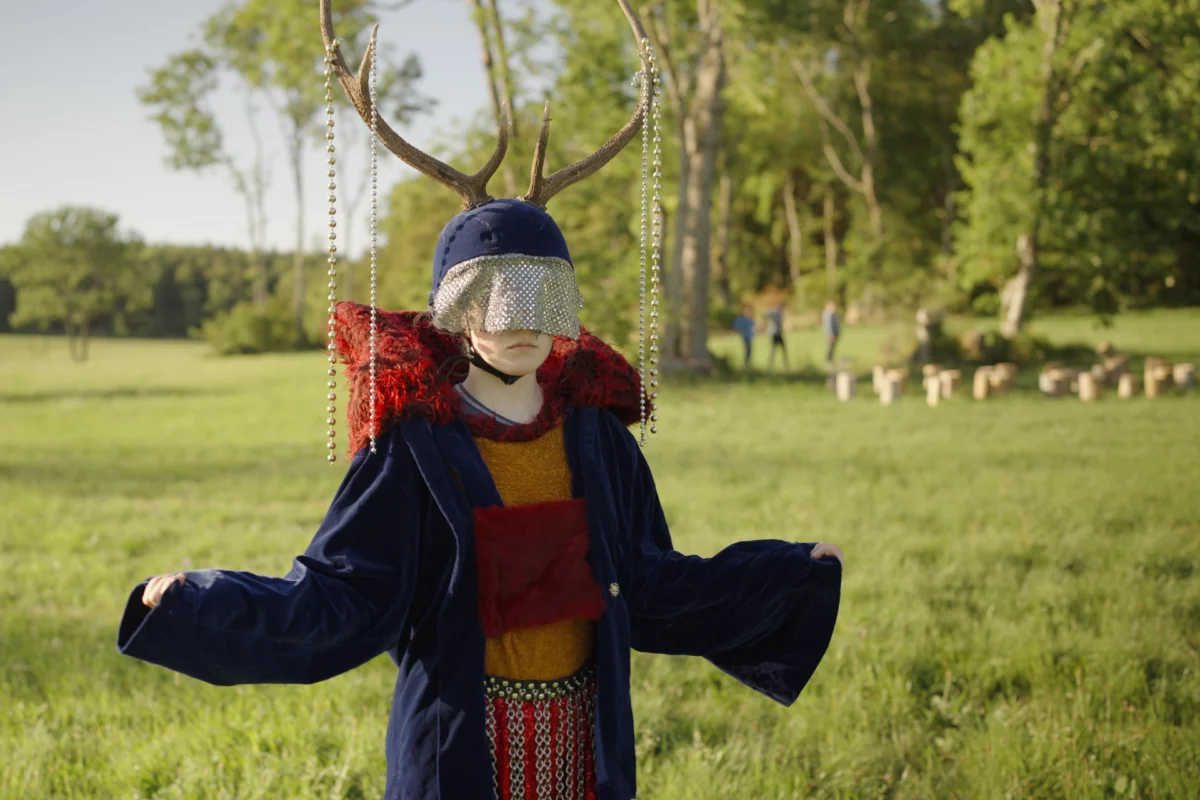 A performer stands in a dark blue coat with a red fur hood on a green field. On her head, the person is wearing a kind of helmet with antlers from which strings of pearls hang. The eyes and nose are covered by a piece of glittering fabric that forms the visor of the helmet.