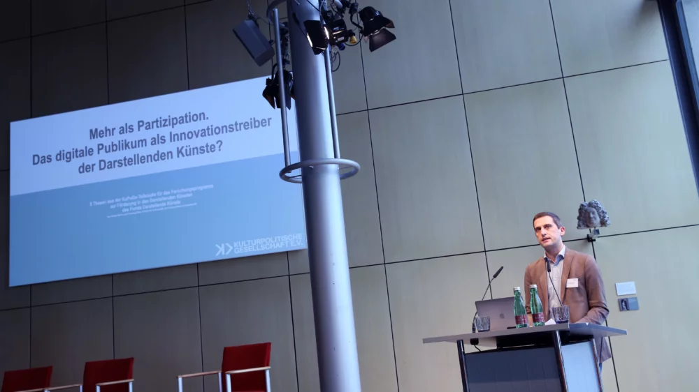 Portrait von Dr. Henning Mohr am Redner*innenpult während des Vortrags. Im Hintergrund ist eine illustrierende Projektion zu sehen.
