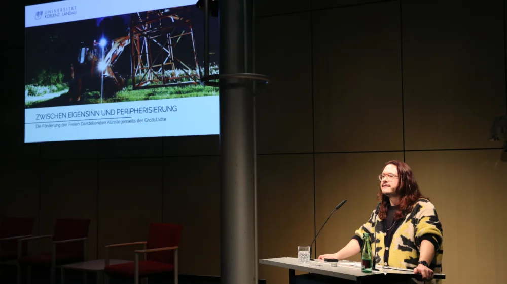 Portrait von Micha Kranixfeld am Redner*innenpult während des Vortrags. Im Hintergrund ist eine illustrierende Projektion zu sehen.