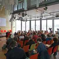Blick in den Saal des Symposiums: Prof. Dr. Thomas Schmidt ist für seinen Vortrag auf einer großen Leinwand zugeschaltet. Das Publikum sitzt in mehreren Stuhlreihen davor.
