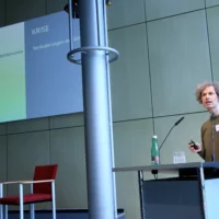 Portrait von Prof. Dr. Kai van Eikels am Redner*innenpult während des Vortrags. Im Hintergrund ist eine illustrierende Projektion zu sehen.