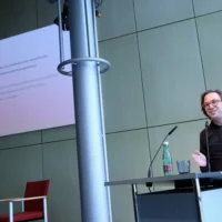Portrait von Dr. Philipp Schulte am Redner*innenpult während des Vortrags. Im Hintergrund ist eine illustrierende Projektion zu sehen.