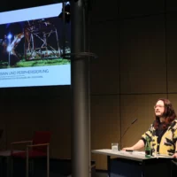 Portrait von Micha Kranixfeld am Redner*innenpult während des Vortrags. Im Hintergrund ist eine illustrierende Projektion zu sehen.