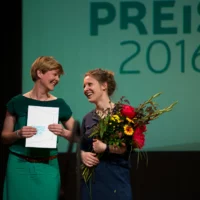 The two members of Pulk Fiktion stand on stage smiling at each other. One holds a bouquet of flowers, the other a certificate in her hands.