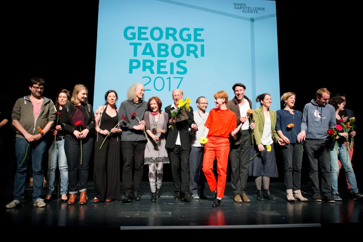 In einer Reihe, mit Blumen in ihren Händen, stehen rund 15 Menschen in einer Reihe und blicken lachend in das Publikum. Im Hintergrund ist der Schriftzug Tabori Preis 2017 auf einer Leinwand zu lesen.