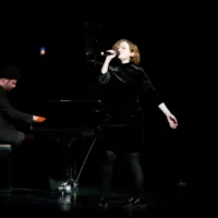 In a black dress, the microphone held in front of her mouth, a singer performs a song. Behind her is a pianist at a concert grand piano.