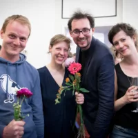 Die vier Preisträger*innen von markus & markus lächelm während der Aftershow-Party in die Kamera, mit Blumen und Sektgläsern in den Händen.