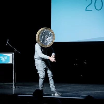 A performer stands on stage in a slightly bent posture. His face is covered by an oversized 1-euro coin that he wears above his head.