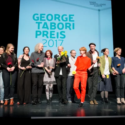 Around 15 people stand in a row, with flowers in their hands, and look into the audience, laughing. In the background, the words Tabori Prize 2017 can be read on a screen.
