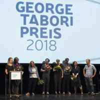 Aufgereiht , zum Teil mit Blumensträußen in den Händen, stehen die Mitglieder von Turbo Pascal nebeneinander auf der Bühne vor großer Leinwand mit dem Tabori-Emblem. Eines von ihnen hält eine Dankesrede am Redner*innenpult und schaut in das Publikum.
