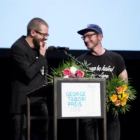 Steffen Klewar und Jörg Albrecht von copy & waste während ihrer Dankesrede auf der Bühne des HAU 1, mit Blumen in den Händen.