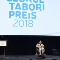 Kübra Sekin, sitting in a wheelchair, moderates the evening on the stage of HAU 1 and addresses her words to the audience.
