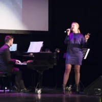 A pianist plays on the concert grand piano. Next to him is a singer in a purple dress. Her eyes closed, she sings powerfully into the microphone.
