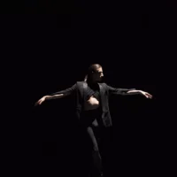 In the spotlight, a black-clad dancer with arms outstretched performs a dance movement on the otherwise dark stage.