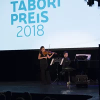 In front of the large screen with the Tabori Prize emblem, a pianist sits playing the grand piano. Next to him, a violinist is engrossed in her playing.