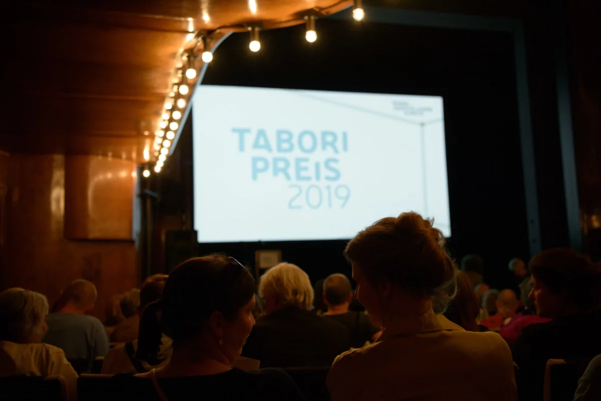 Im abgedunkelten Zuschauerraum des HAU 1 schauen Menschen auf die erleuchtete Leinwand mit dem Schriftzug "Tabori Preis 2019" auf der Bühne.