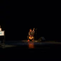 Drei Menschen stehen vereinzelt in Spotlights auf der sonst dunklen Bühne des HAU 1. Im Zentrum sitzt eine Musikerin und spielt das Cello, links und rechts von mir tragen die Personen abwechselnd einen Text vor.
