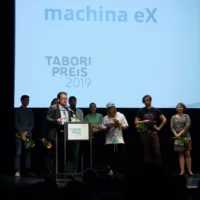 Die Performer*innen von machina eX bei der Preisübergabe auf der Bühne des HAU 1. Robin Hädicke steht für seine Dankesrede am Mikrofon des Redner*innenpults.