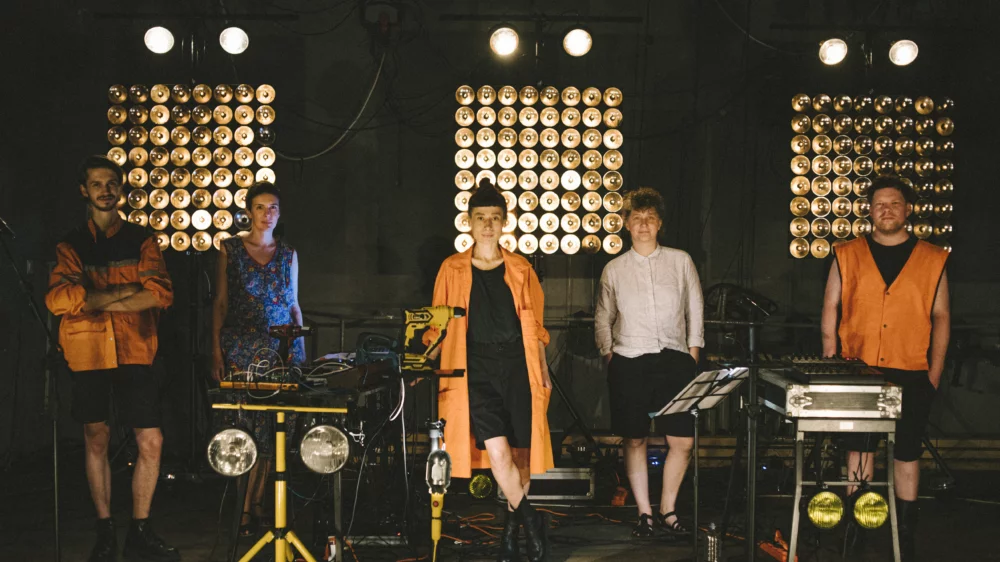 Scene from a performance by Teatru Spalatorie: five performers stand behind stage structures that resemble musical installations and look into the camera. In the background, dim spotlights arranged in three square fields can be seen, directed at the audience.