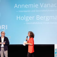 Holger Bergmann und Annemie Vanackere eröffnen die Veranstaltung vor großer Leinwand auf der Bühne des HAU 1.