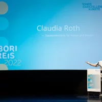 Kulturstaatsministerin Claudia Roth hält am Redner*innenpult mit weit ausgebreiteten Armen ihre Rede zur Eröffnung des Tabori Preises vor großer Leinwand.