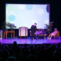 Christiane Rösinger bei ihrem musikalischen Beitrag auf der Bühne des HAU 1. Im Hintergrund sieht man die mit Tierköpfen kostümeriten Performer*innen in einer Sitzlounge.