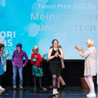 Minister of State for Culture Claudia Roth stands on stage and applauds the members of "Meine Damen und Herren" who are also on stage. One member holds the trophy of the Tabori Prize in her hands.