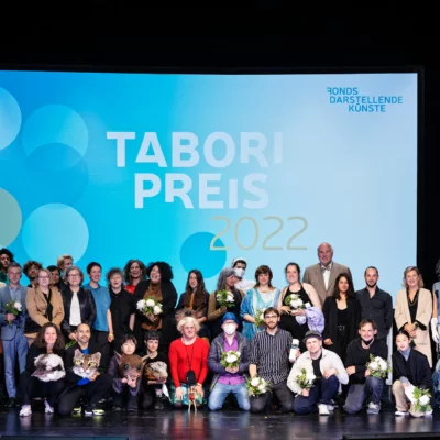 In drei Reihen versammeln sich die diesjährigen Preisträger*innen und Programmbeteiligten auf der Bühne des HAU 1 zum Abschlussfoto der Preisverleihung. Im Hintergrund ist der Schriftzug Tabroi Preis 2022 auf großer Leinwand zu lesen.