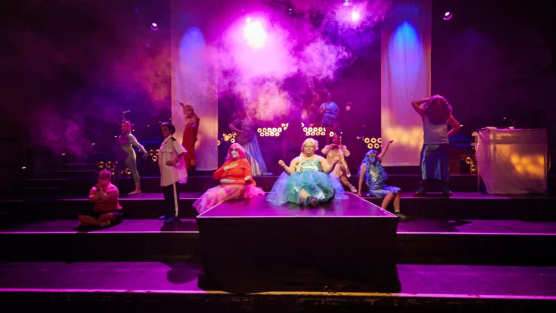 Zwölf Performer*innen des Ensembles "Meine Damen und Herren" sind in bunten Kostümen auf der Bühne verteilt, in buntes Licht getaucht. Von der Decke senkt sich Nebel herab.