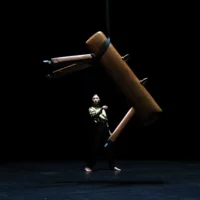 A pommel horse hangs on a rope in front of a dancer who is illuminated from above.