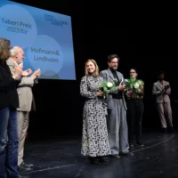 Die Preisträger*innen Hofmann und Lindholm mit Blumen in der Hand in der MItte der Bühne. Seitlich von ihnen applaudieren ihnen die Redner*innen.