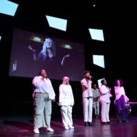 Five people dressed in white with microphones in their hands, some of them singing. In the background, Lisa Lucassen can be seen on a screen as the conductor.