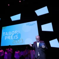 Holger Bergmann speaks to the audience from the stage. Behind him, in the dimly pink-lit stage, five people dressed in white and a large screen with the lettering of the Tabori Prize can be seen.
