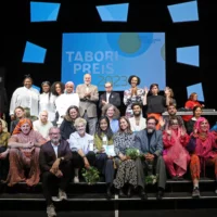 Gruppenbild mit den rund 30 Teilnehmer*innen des Abends auf den Stufen der HAU1-Bühne. Im Hintergrund eine große Leinwand mit dem Tabori Preis-Schriftzug.