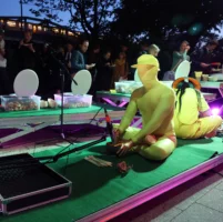 Two performers sit cross-legged on a platform covered with green artificial turf. One of them operates a kind of sound bowl, above which a microphone is attached. In the background, in the semi-darkness under the open sky in front of the HAU 2 people are standing at the buffet.