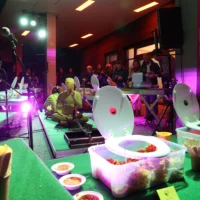 Around two performers in yellow full-body suits in front of HAU 2, the buffet is set up on tables covered with artificial turf. Large plastic boxes with the food are covered with unfolded plastic toilet lids. People help themselves to the buffet in the background.