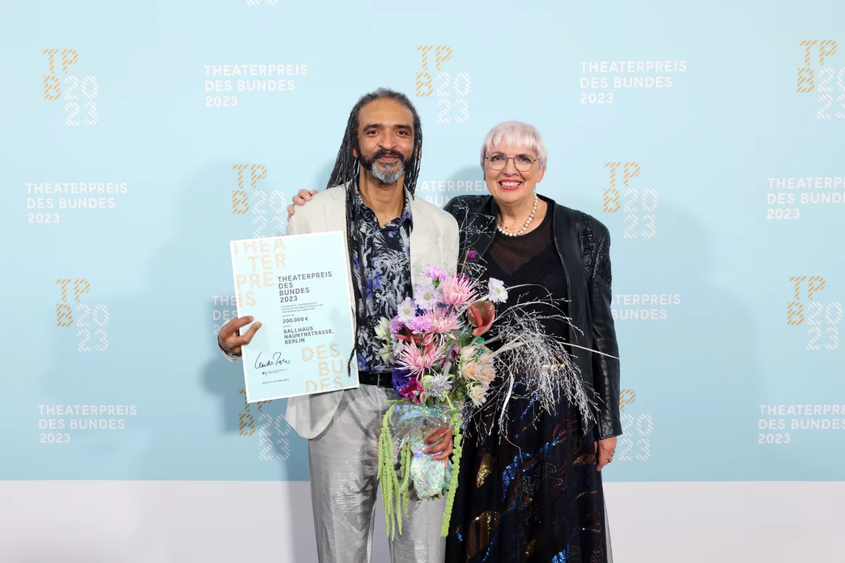 Wagner Carvalho (Artistic Director and Management Ballhaus Naunynstraße) with Minister of State for Culture Claudia Roth. Wagner Carvalho holds a certificate in his right hand and a bouquet of flowers in his left.