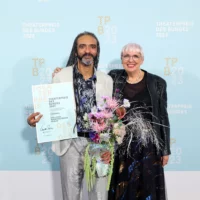 Wagner Carvalho (Artistic Director and Management Ballhaus Naunynstraße) with Minister of State for Culture Claudia Roth. Wagner Carvalho holds a certificate in his right hand and a bouquet of flowers in his left.