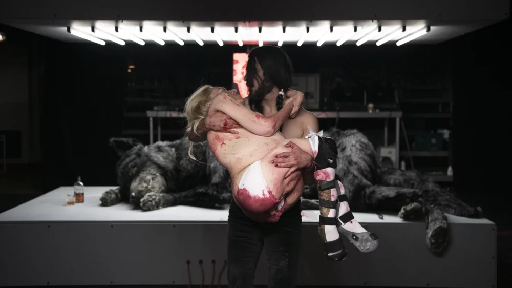 One person stands in the foreground, holding a second blood-covered person in his arms. In the background, a wolf-like animal lies on a treatment table under a lamp made of neon tubes.