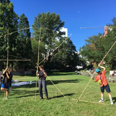 Three people stand on a green area and hold long wooden sticks together.