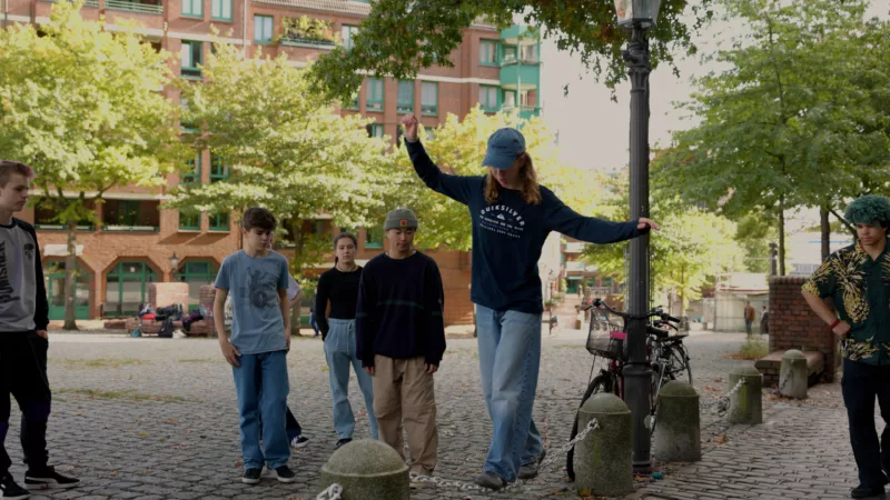 A teenager in a baseball cap balances on a thick metal chain stretched between two concrete bollards. Other young people in the group watch her as she does this.